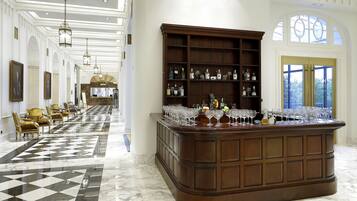 Um hall espaçoso com piso de mármore quadriculado, uma área de bar central com prateleiras de garrafas e copos, e um espaço de estar com poltronas estofadas.