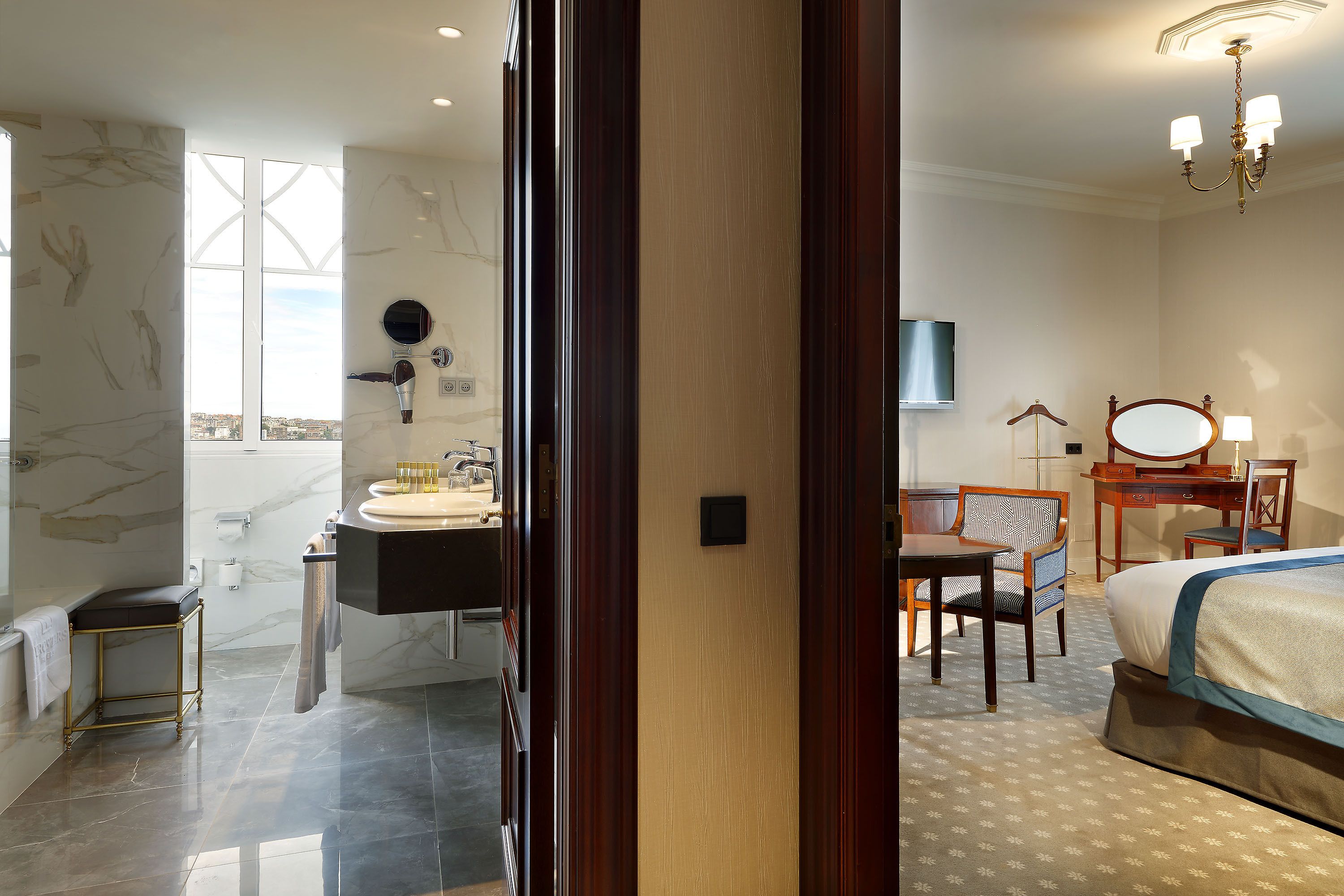 Habitación de hotel con cama, escritorio, silla, tocador con espejo y vistas a la ciudad a través de amplias ventanas.