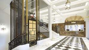 Un amplio vestíbulo con piso a cuadros, una escalera con barandillas ornamentadas y un mostrador de recepción de mármol.