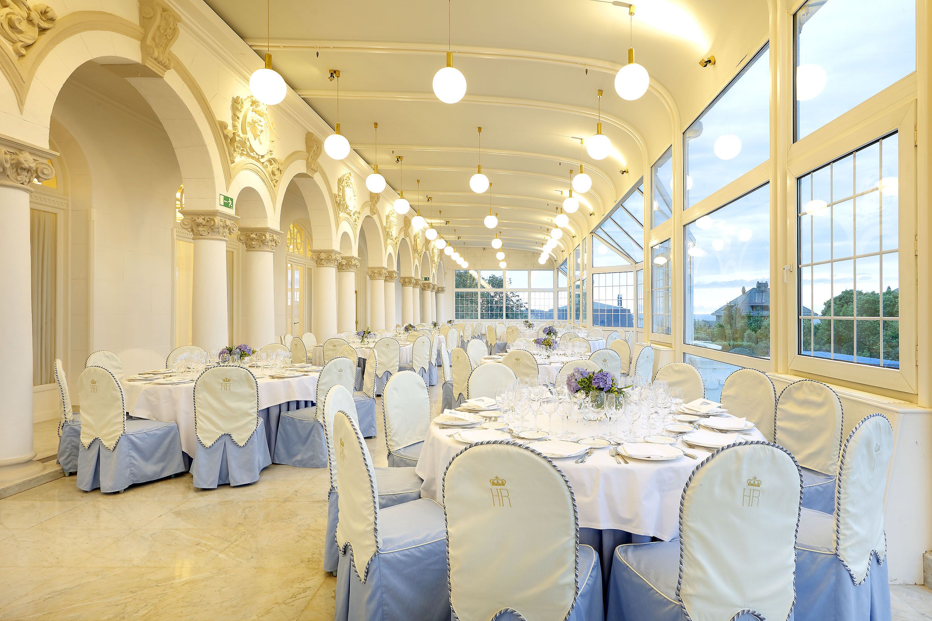Un salón de banquetes con varias mesas redondas, manteles blancos y sillas con protectores de asiento bordados.