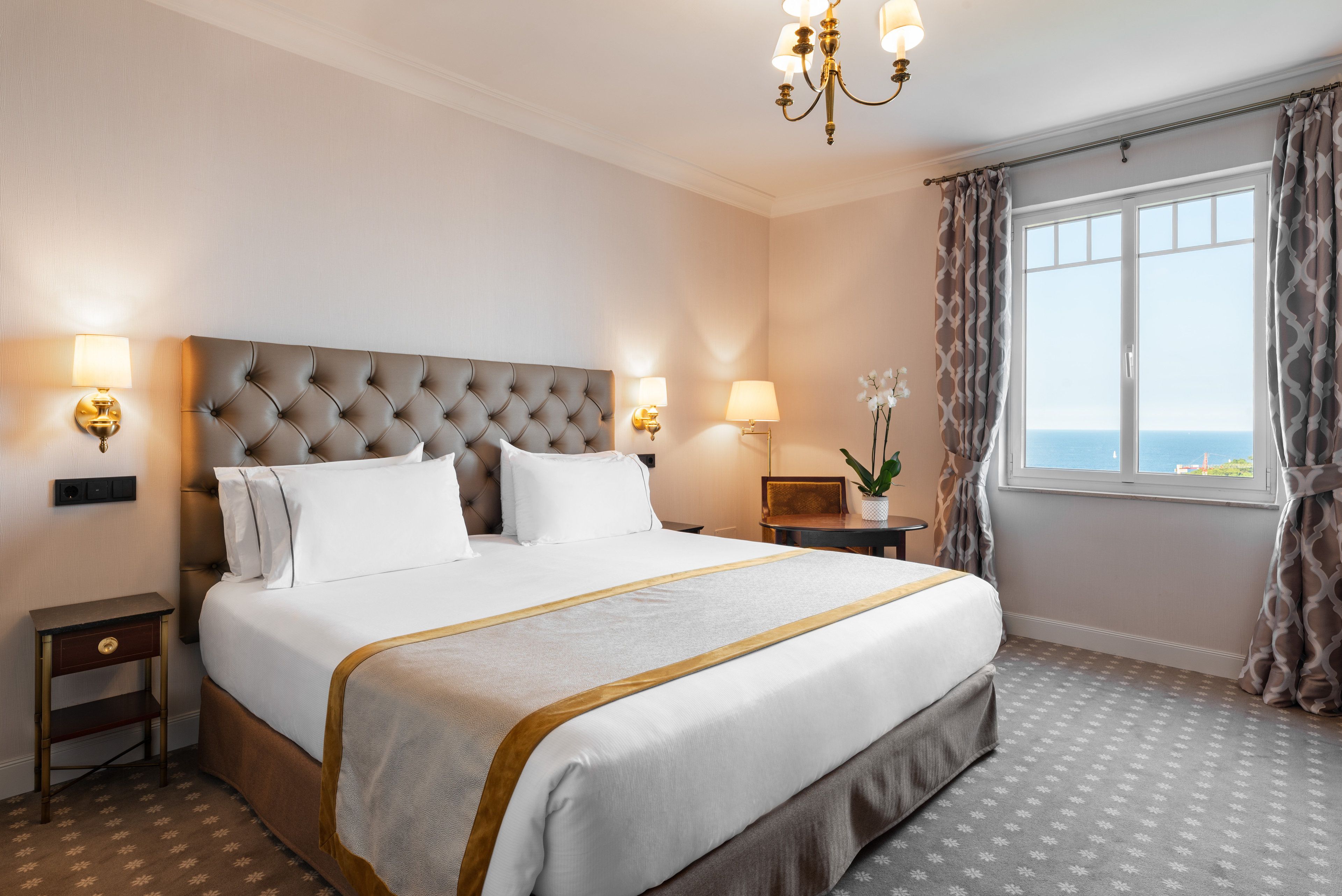 Una habitación de hotel con una cama grande, dos mesitas de noche, un candelabro, una ventana con cortinas y vistas al mar.