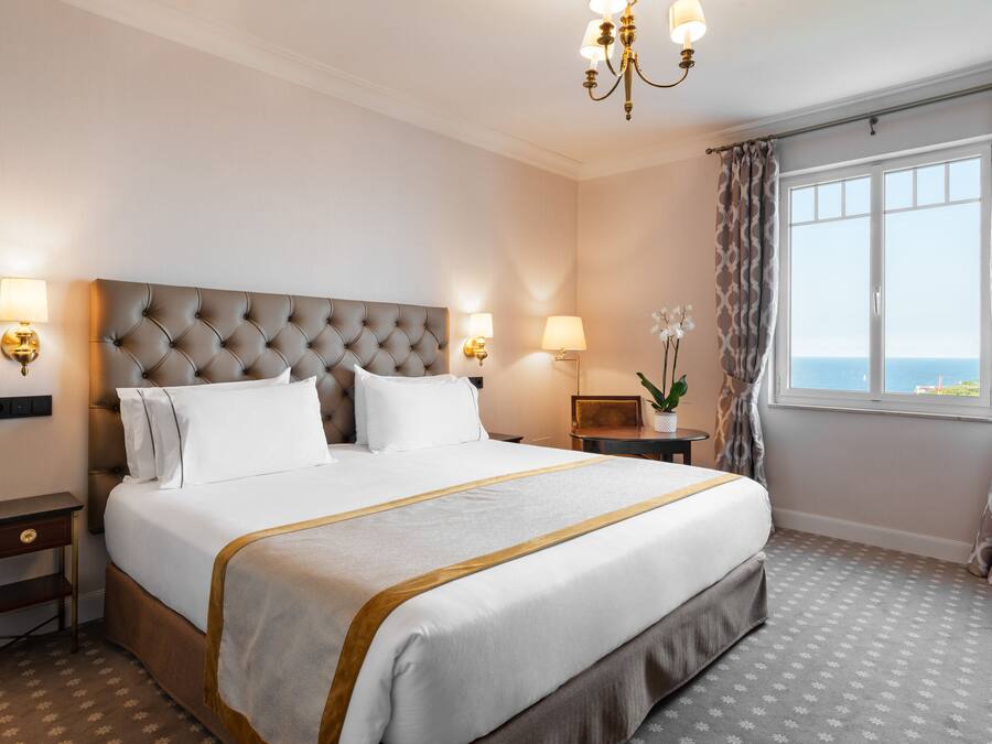 Una habitación de hotel con una cama grande, dos mesitas de noche, un candelabro, una ventana con cortinas y vistas al mar.