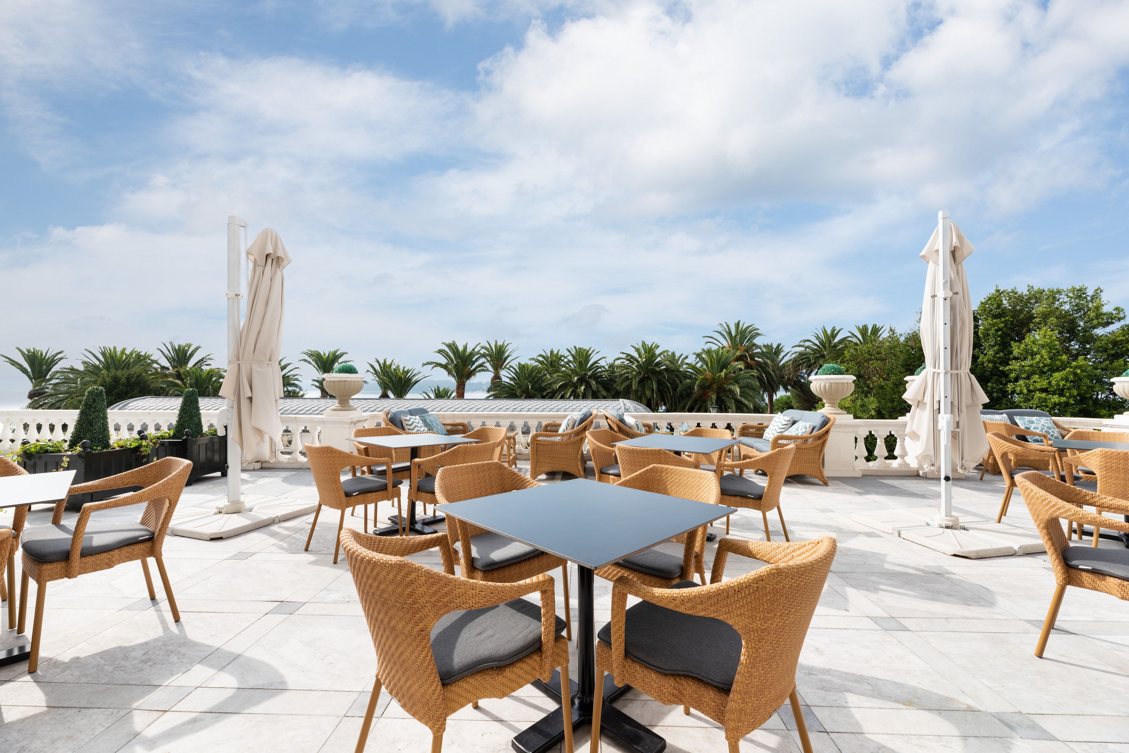 An outdoor patio area with wicker chairs and tables, surrounded by palm trees and a clear sky.