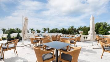 An outdoor patio area with wicker chairs and tables, surrounded by palm trees and a clear sky.