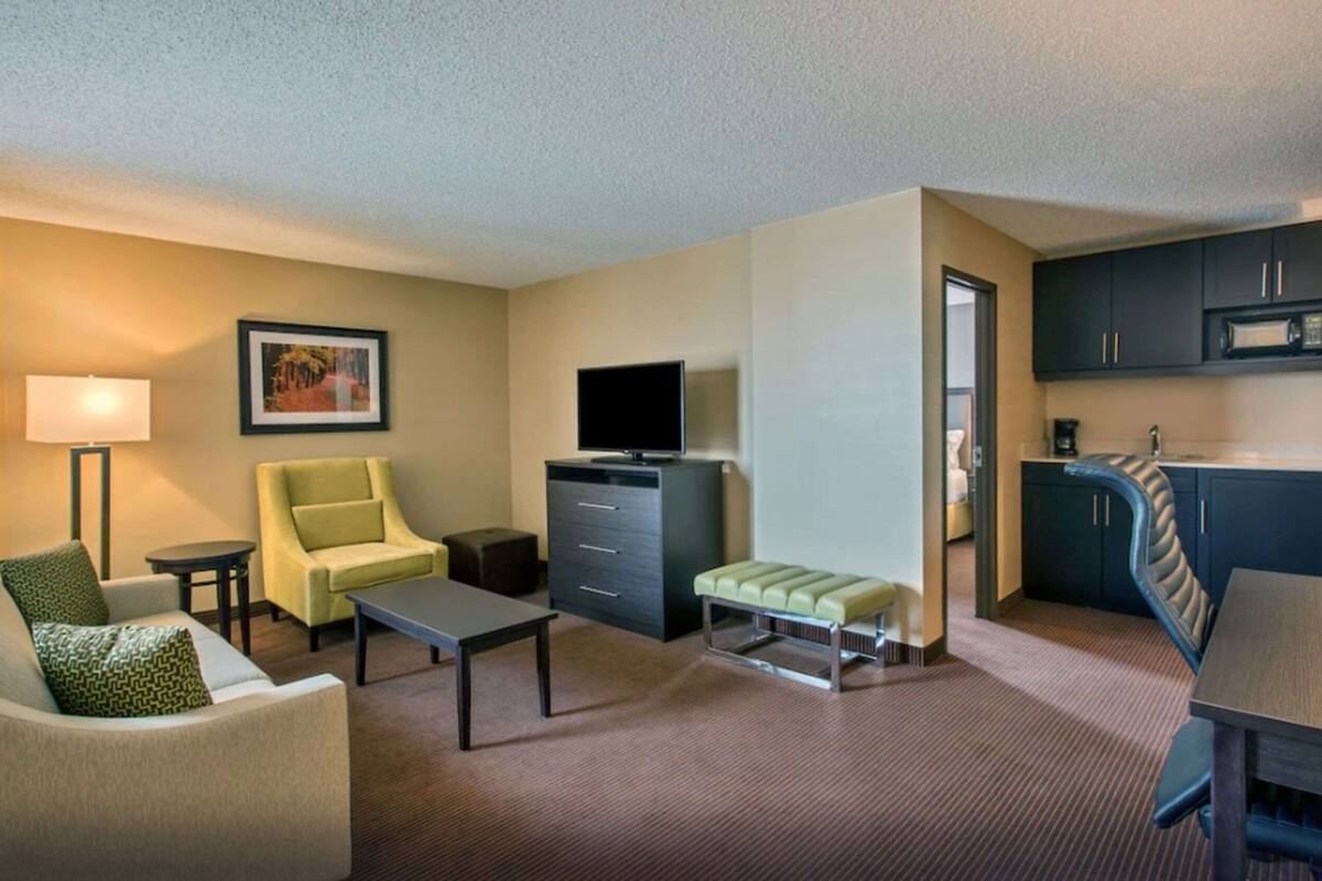 A hotel room with a sofa, armchair, coffee table, TV, kitchenette, and a framed picture on the wall.