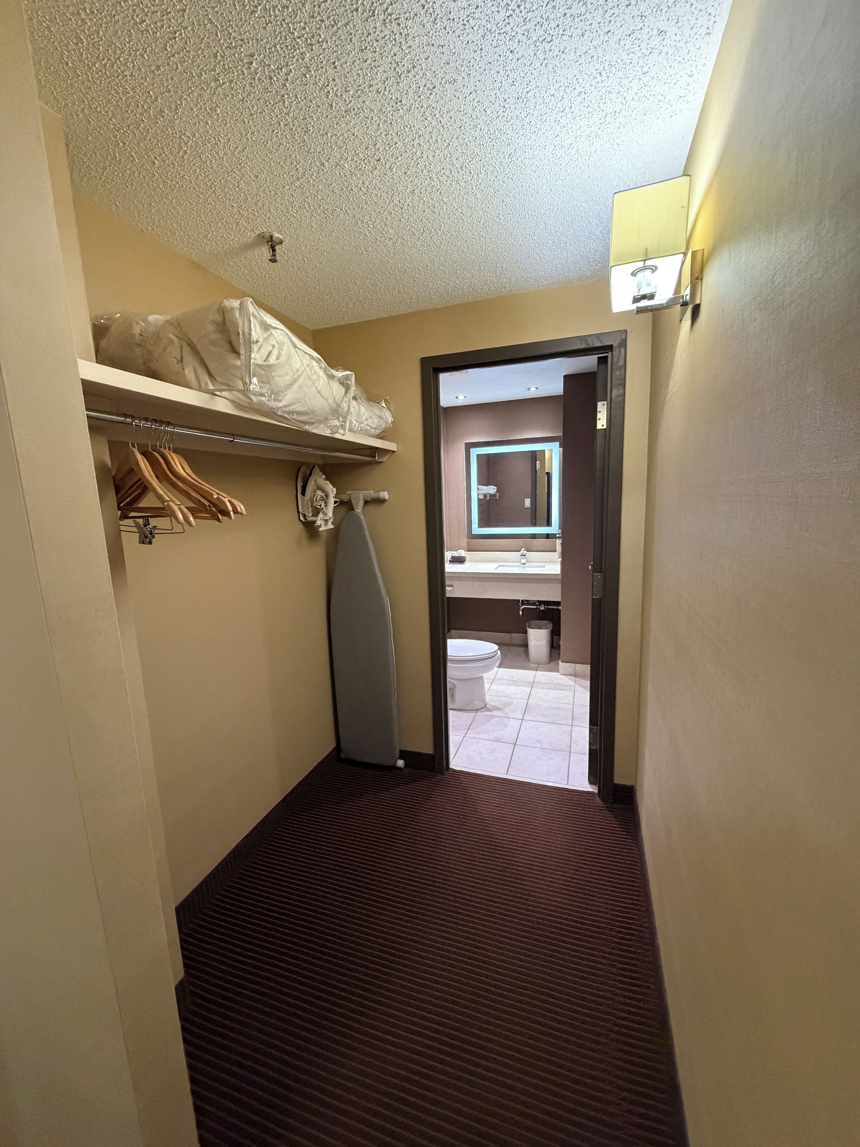 Habitación de hotel con cama, tabla de planchar pequeña, perchero y vista al baño a través de una puerta abierta.