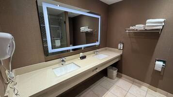Une salle de bain avec un grand miroir, deux lavabos, un sèche-cheveux, un porte-serviettes et une poubelle.