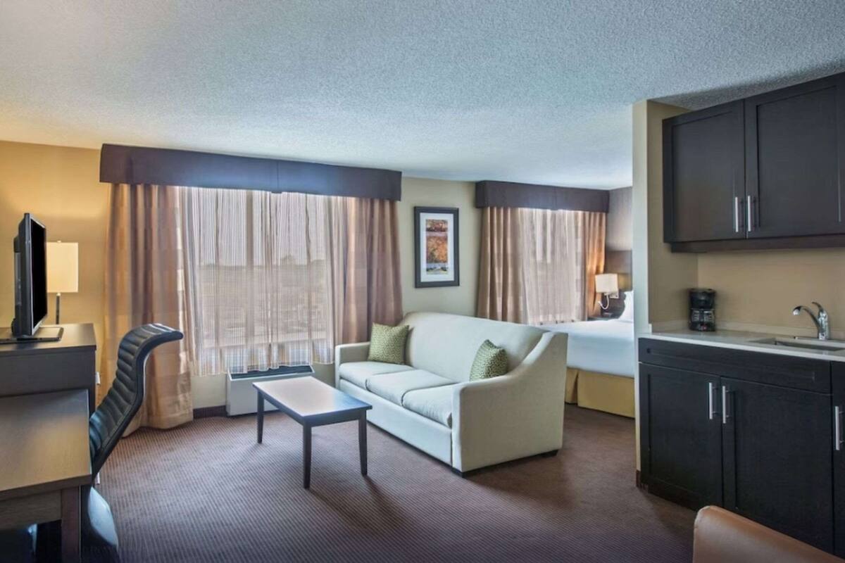 A hotel room with a sofa, a small table, a TV, a desk, a chair, a kitchen area with cabinets, and a window with curtains.