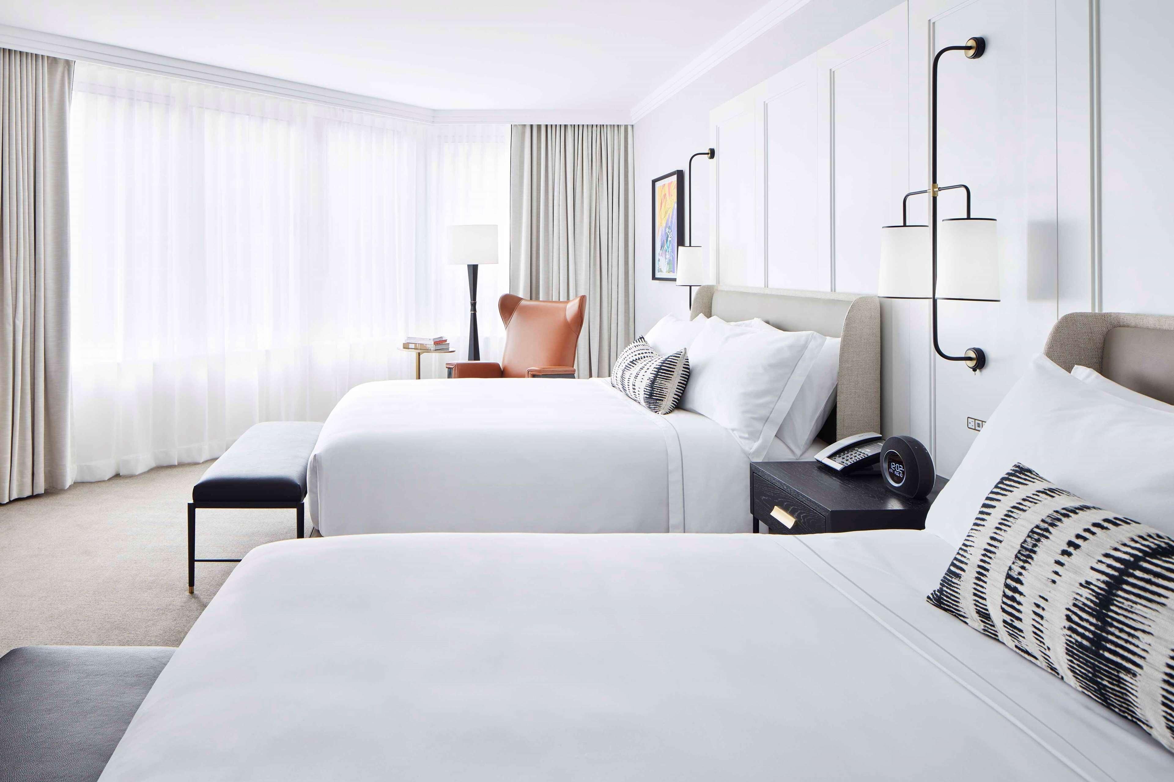 A hotel room with two beds, a chair, a bedside table with a lamp, and a wall-mounted light fixture.