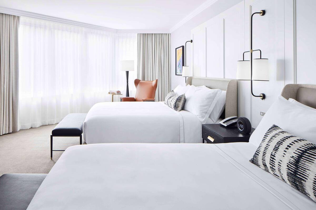 A hotel room with two beds, a chair, a bedside table with a lamp, and a wall-mounted light fixture.