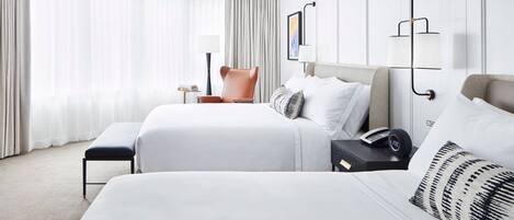 Quarto de hotel com duas camas, uma cadeira, mesa de cabeceira com abajur e luminária na parede.