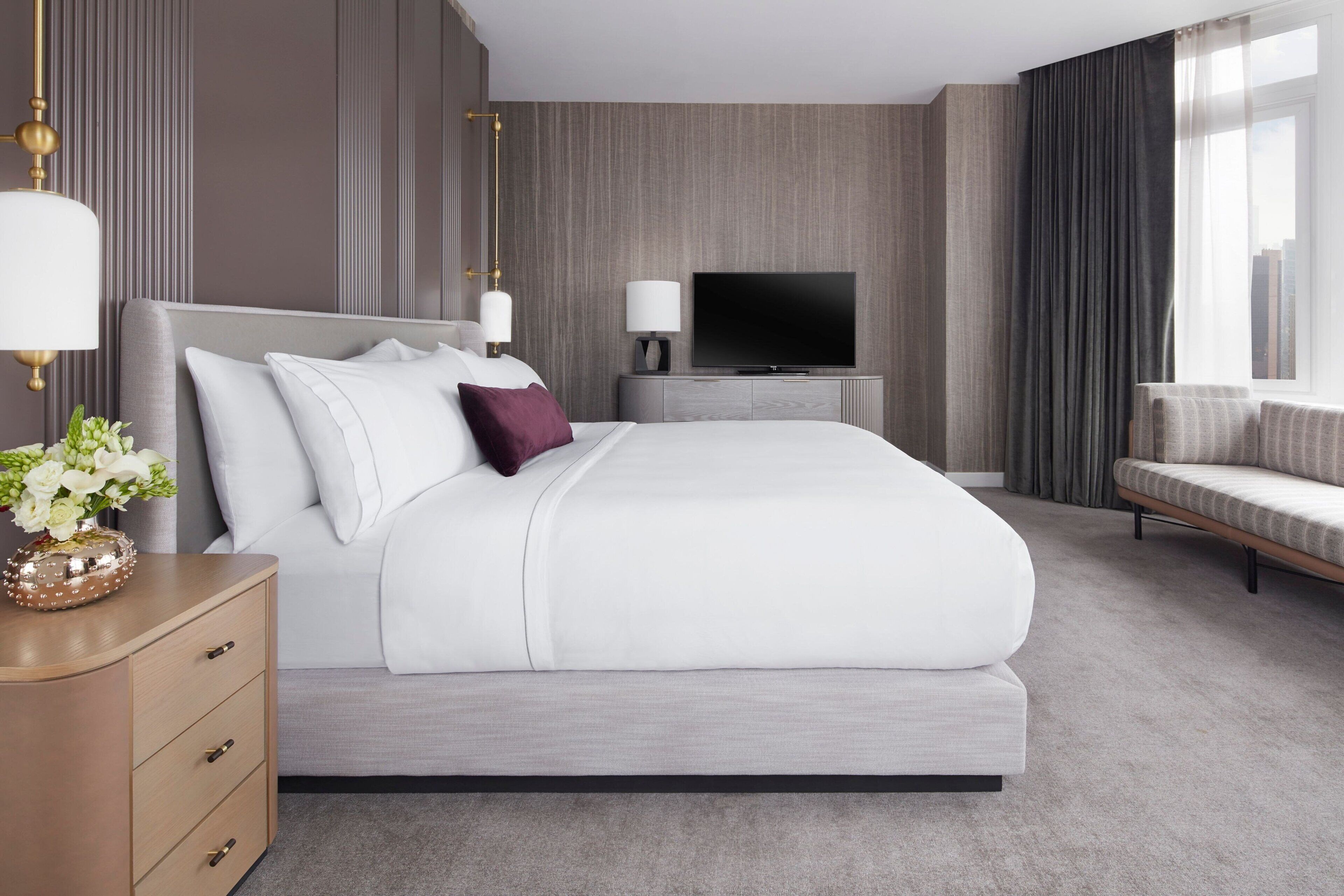 A modern hotel room with a large bed, a TV, a bedside table with a lamp, and a sofa.