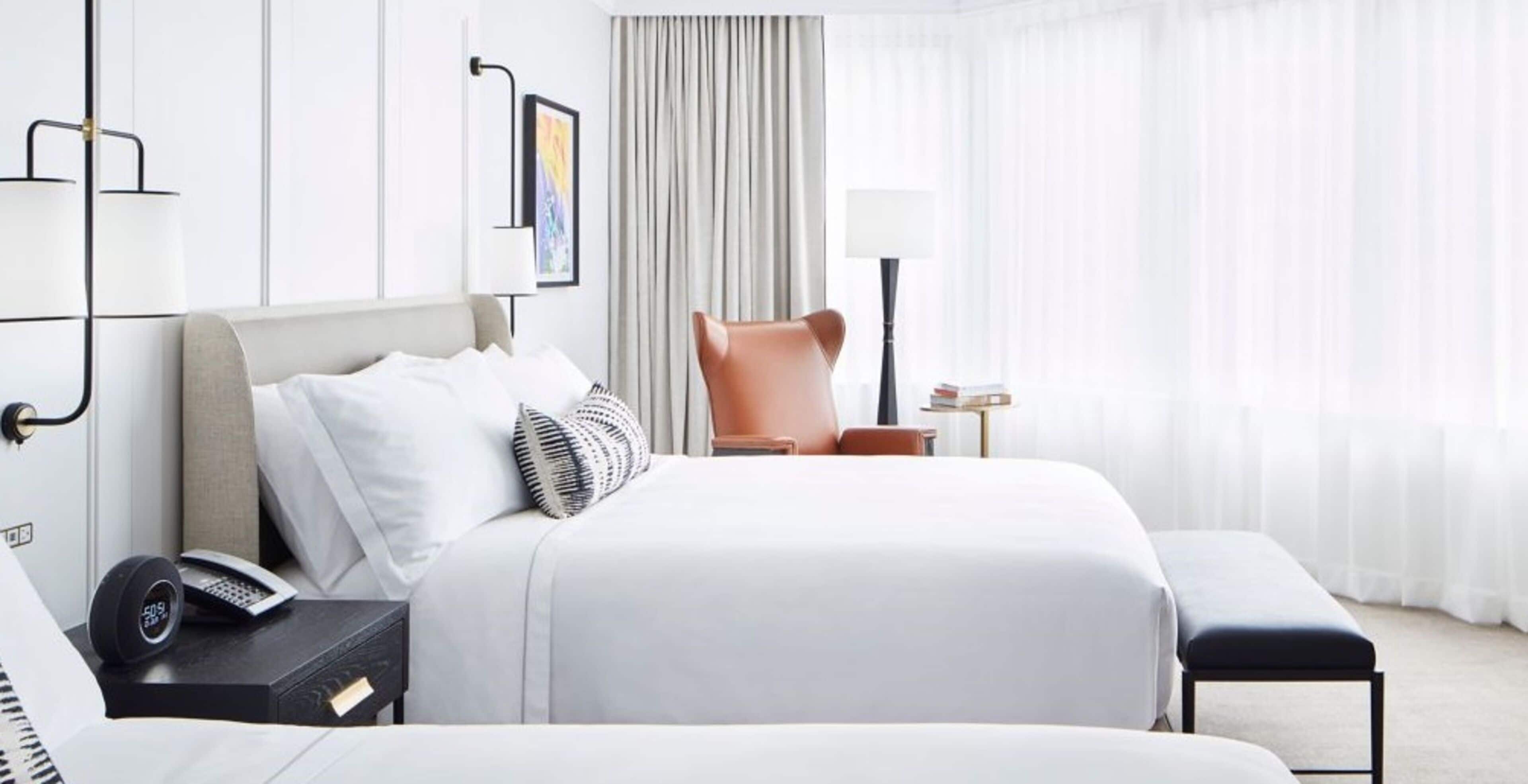 A hotel room with a large bed, a bedside table with a phone and a lamp, a chair, and a window with curtains.
