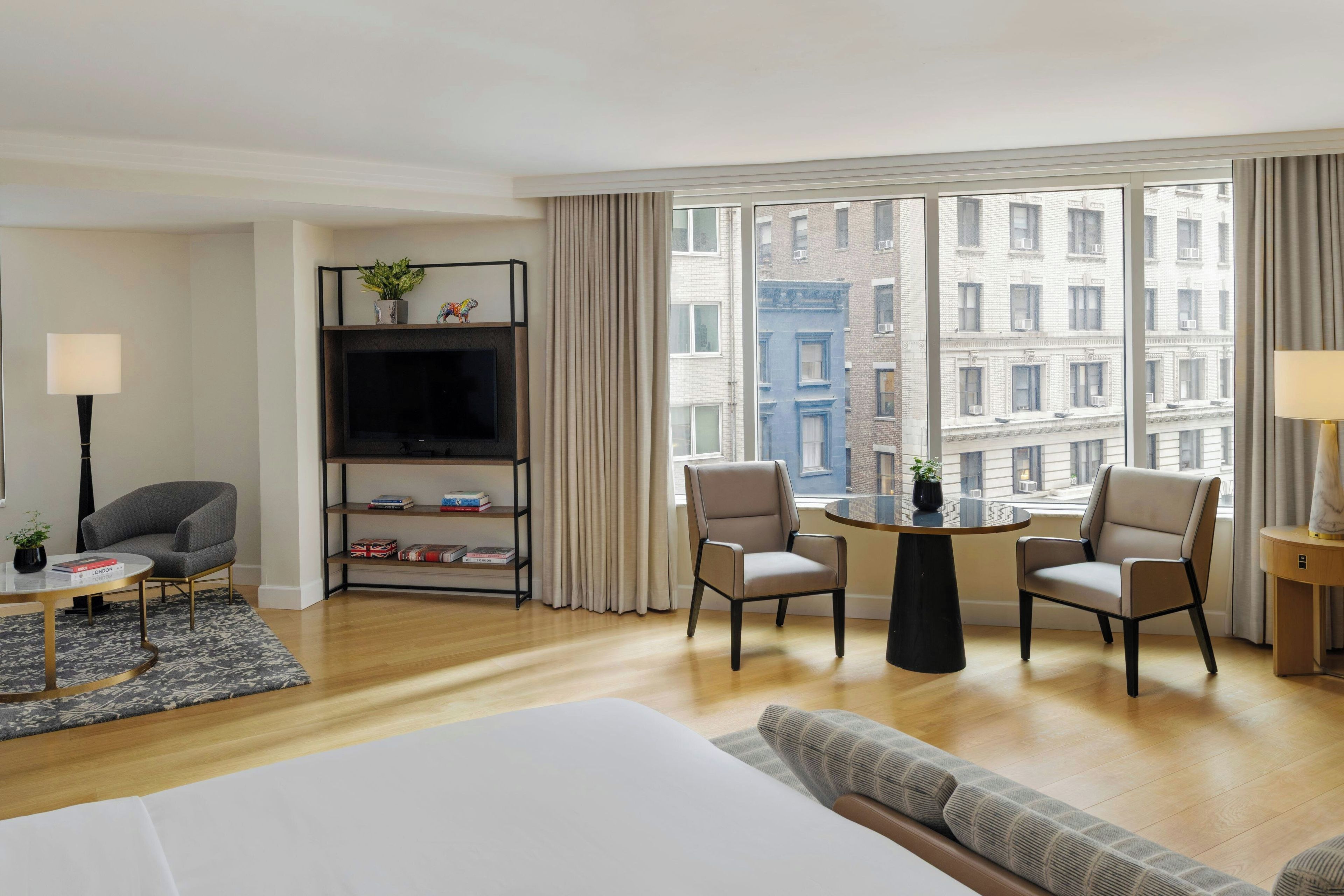 A hotel room with a bed, a TV, a bookshelf, two armchairs, a small table, and a view of buildings through large windows.