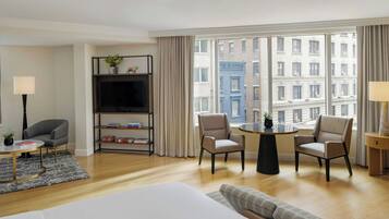 A hotel room with a bed, a TV, a bookshelf, two armchairs, a small table, and a view of buildings through large windows.