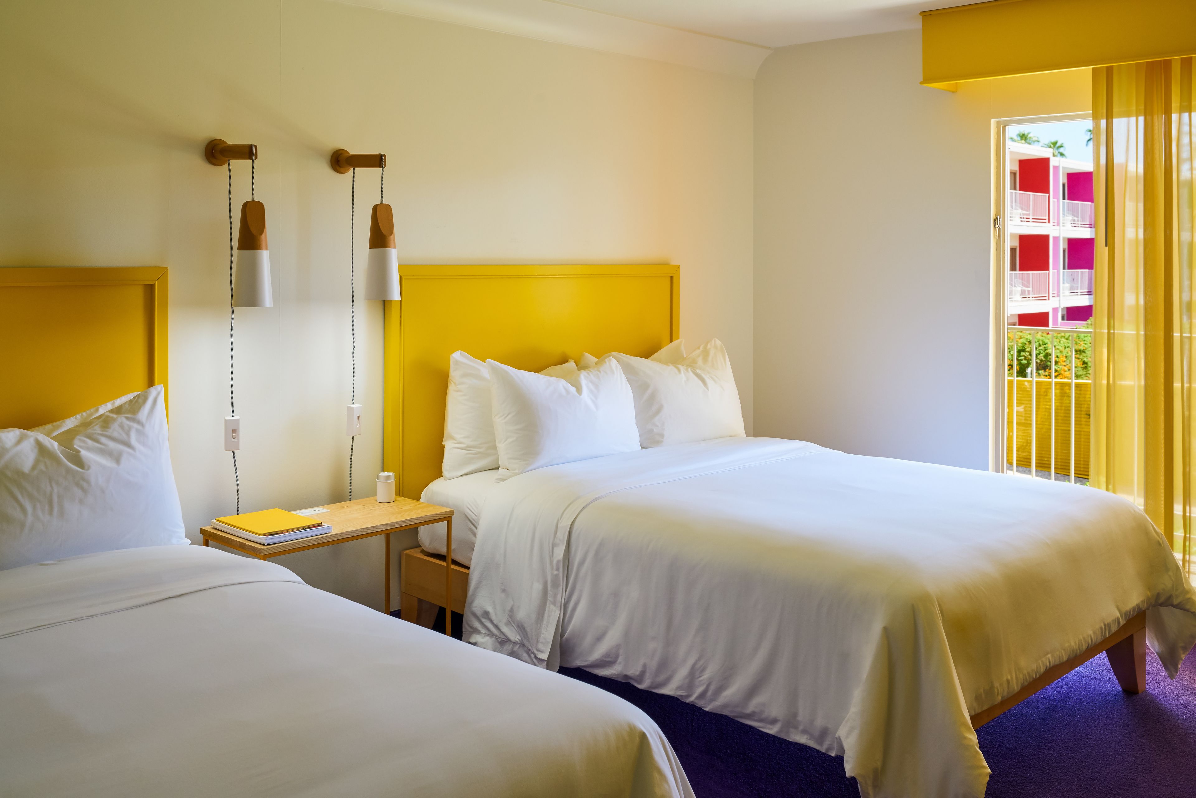 A hotel room with two beds, a nightstand, and a window with yellow curtains.