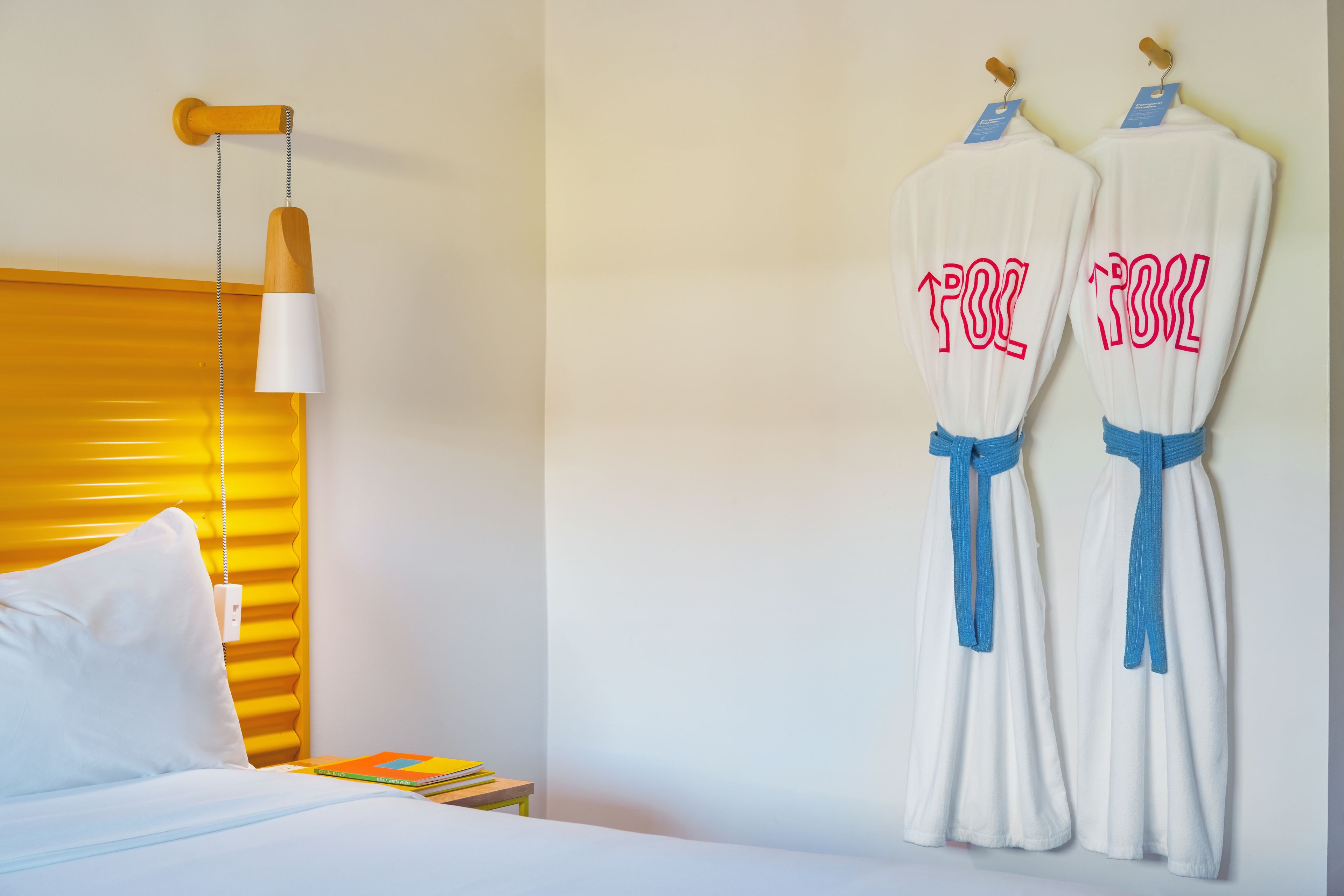 A hotel room with a bed, a bedside table with books, a lamp, and two robes hanging on the wall.