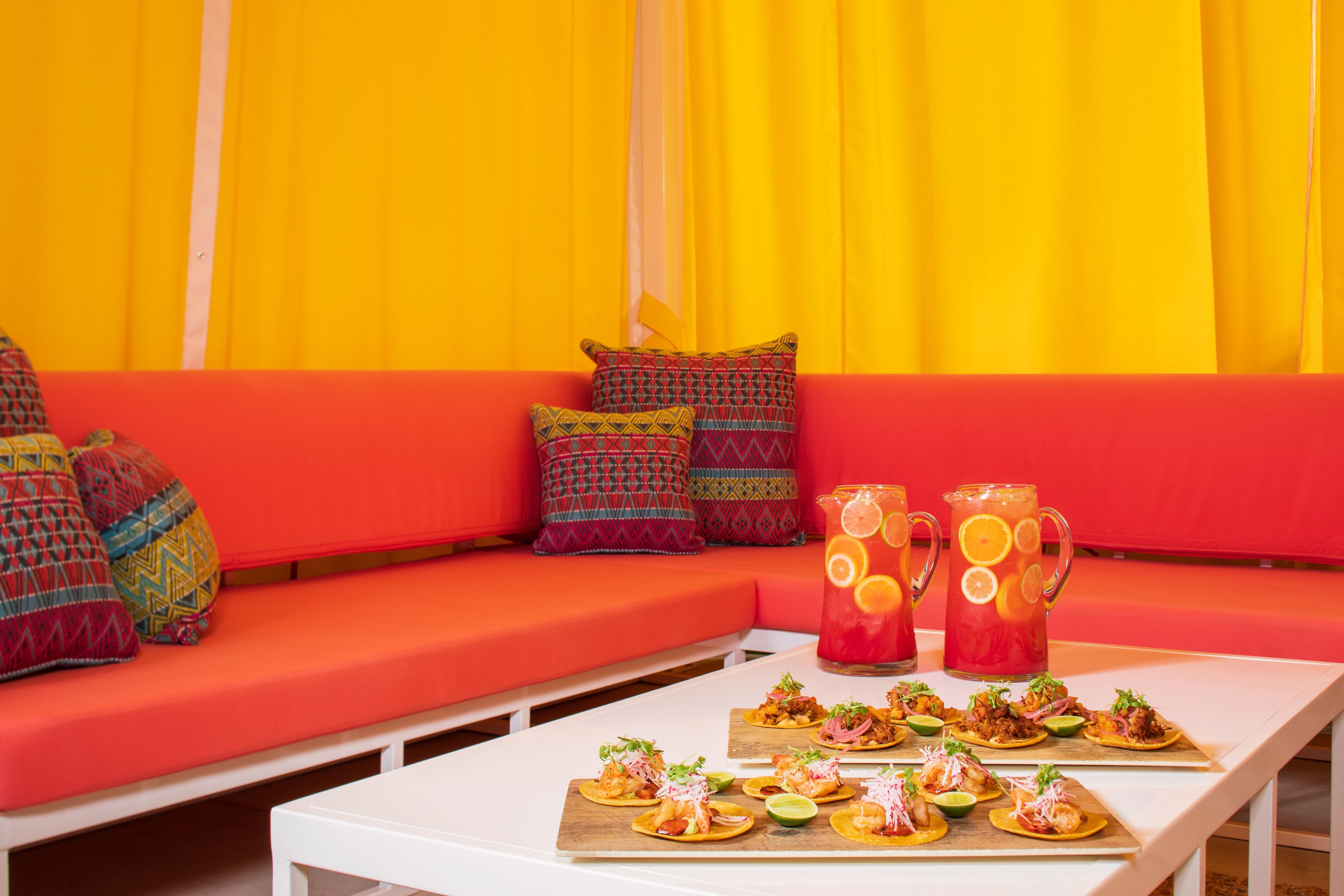A dining area with a white table set with appetizers, a red sofa with patterned cushions, and two pitchers of a red beverage with citrus slices.