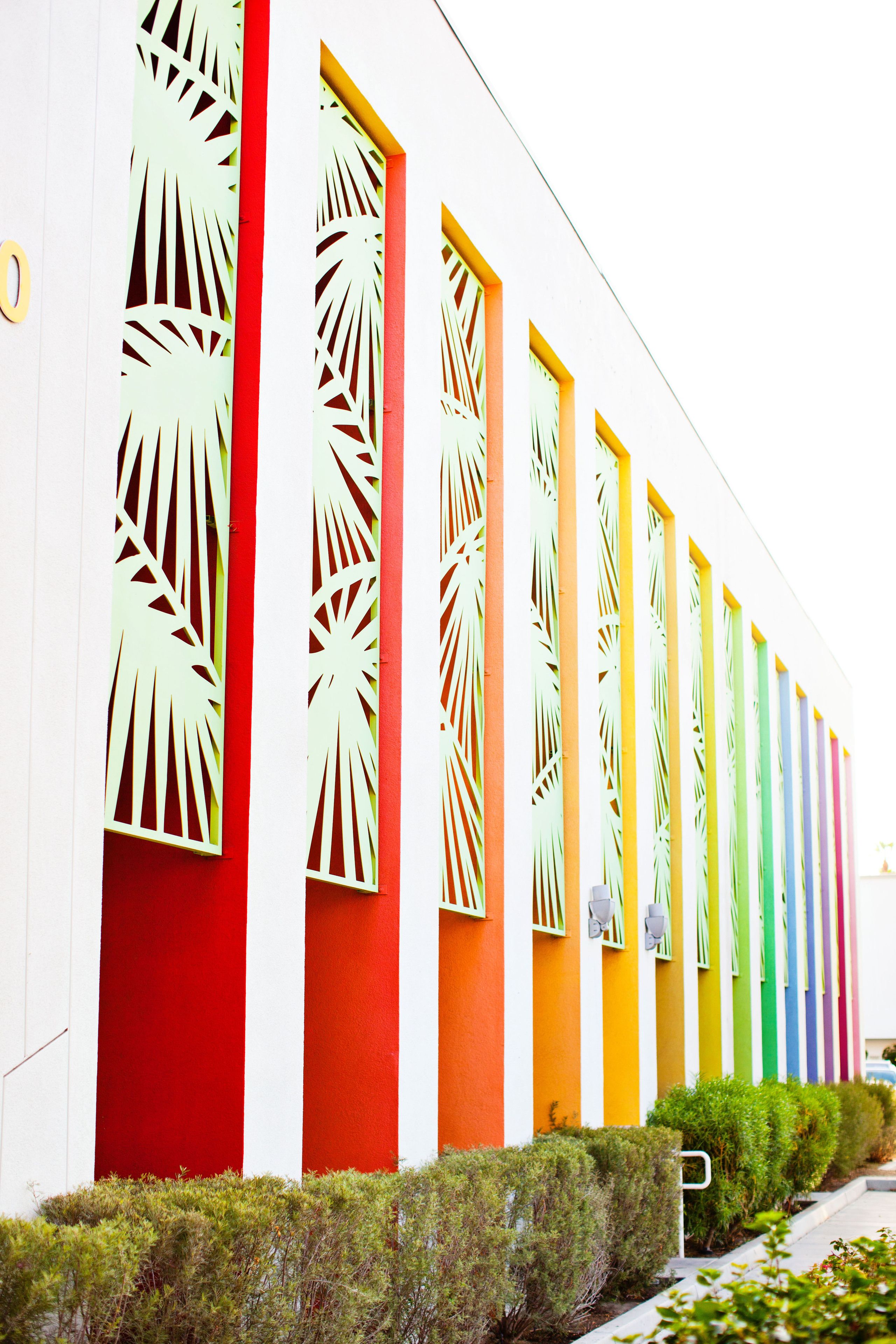 A row of brightly colored doors with geometric cutouts, set against a white building facade.
