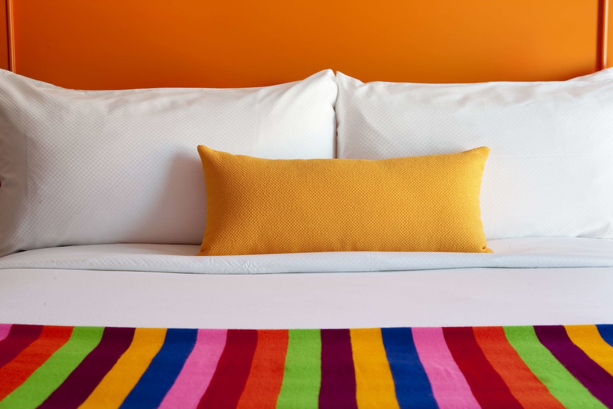 A bed with white pillows and a single yellow pillow, and a rainbow-striped blanket.
