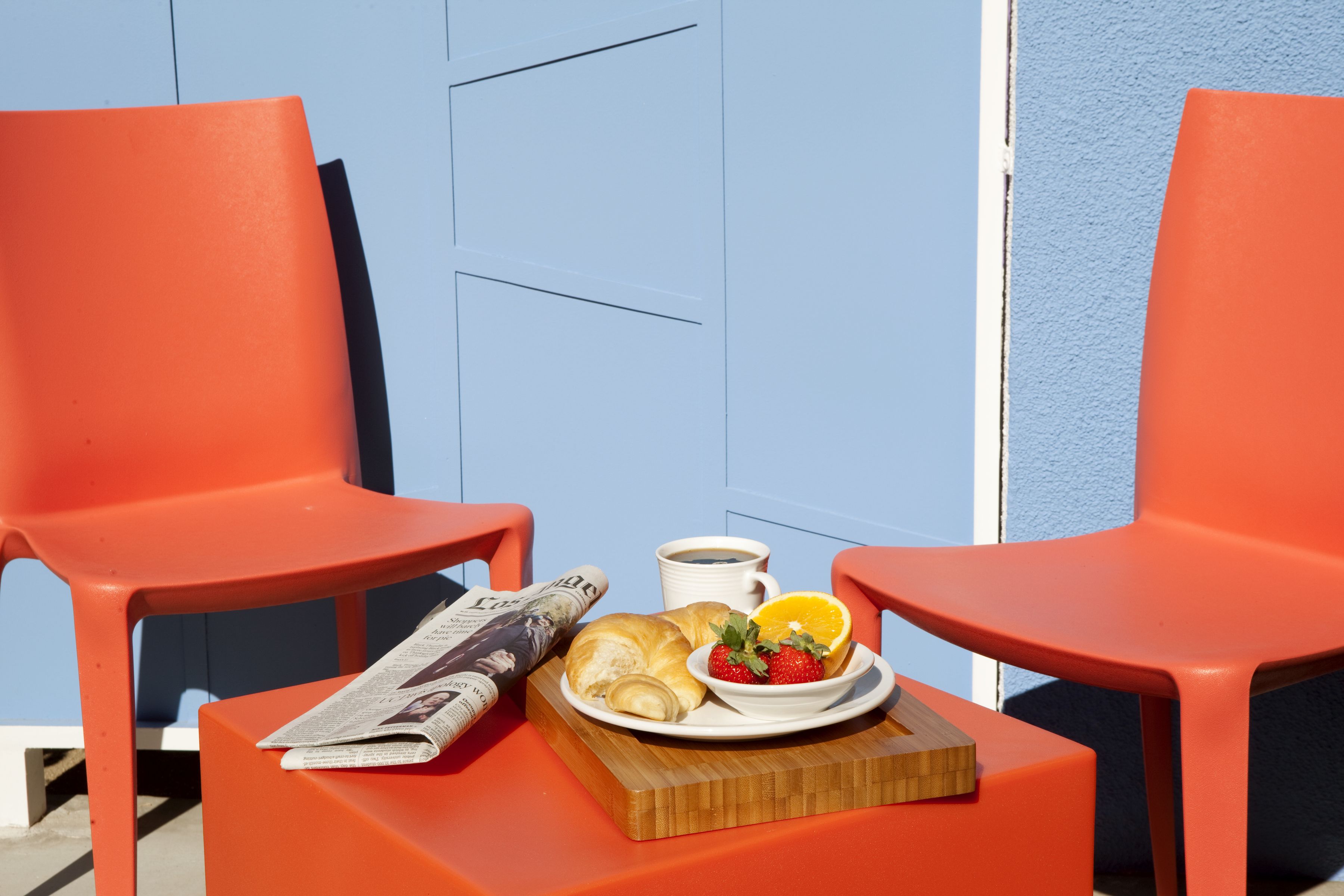 Two orange chairs, a wooden table with a plate of food, a cup of coffee, and a newspaper.