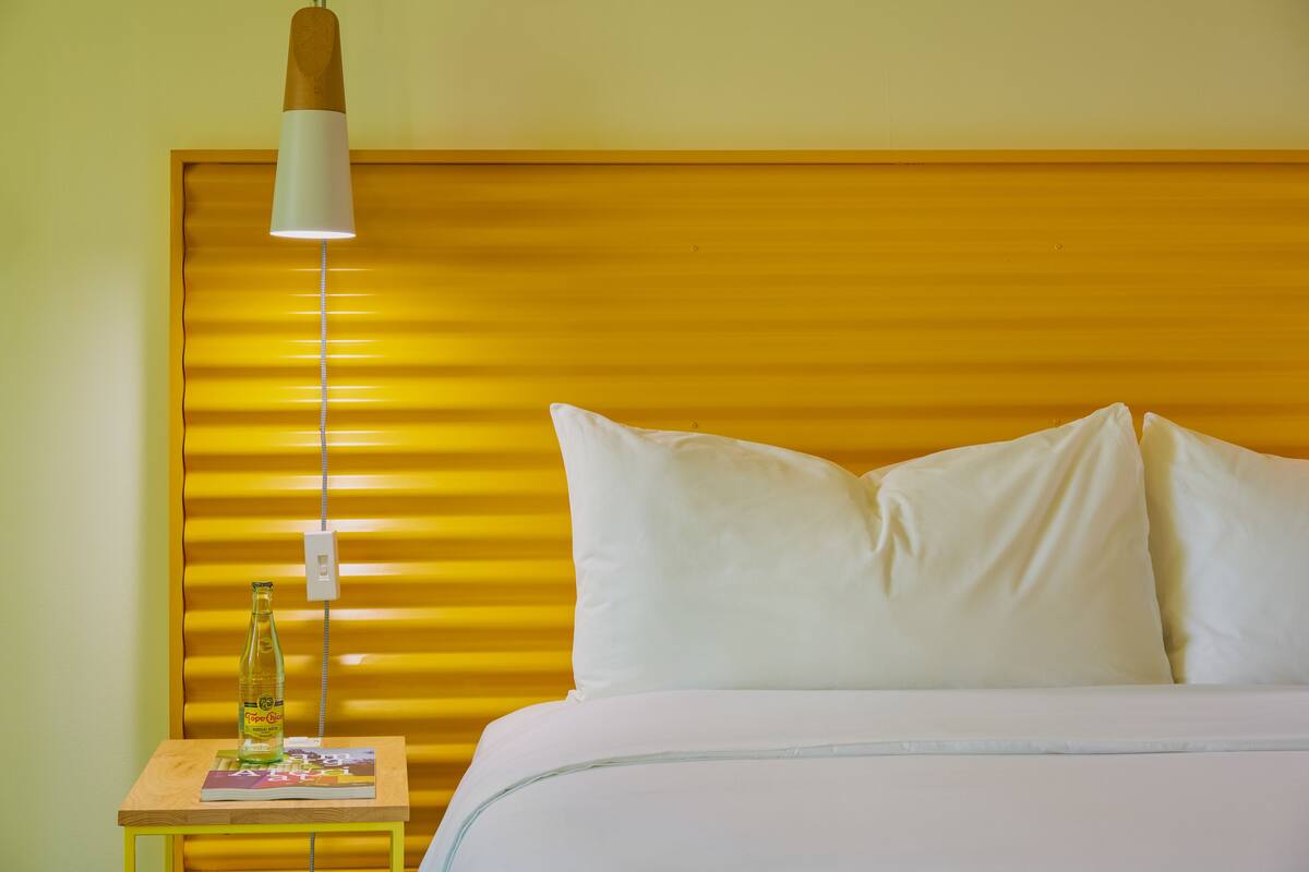 A hotel room with a bed, white pillows, a bedside table with a bottle, and a lamp.