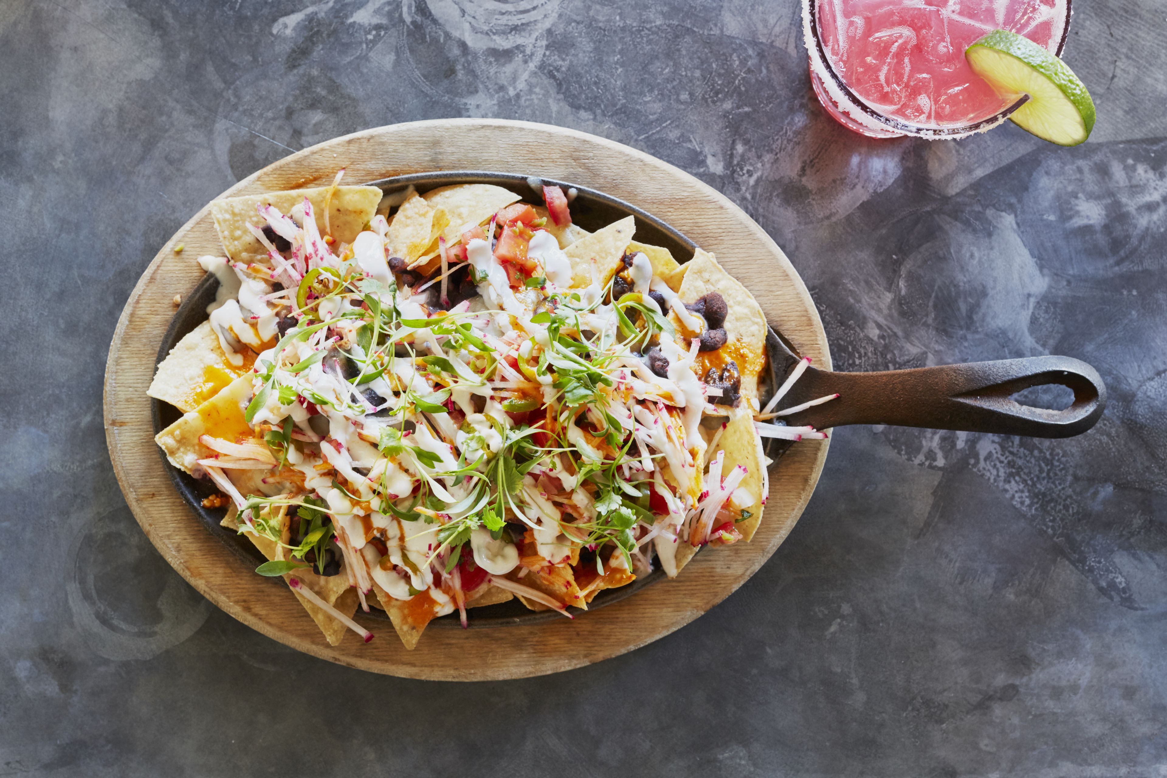A wooden serving board with nachos topped with various ingredients and a drink with a lime wedge.