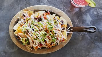 A wooden serving board with nachos topped with various ingredients and a drink with a lime wedge.