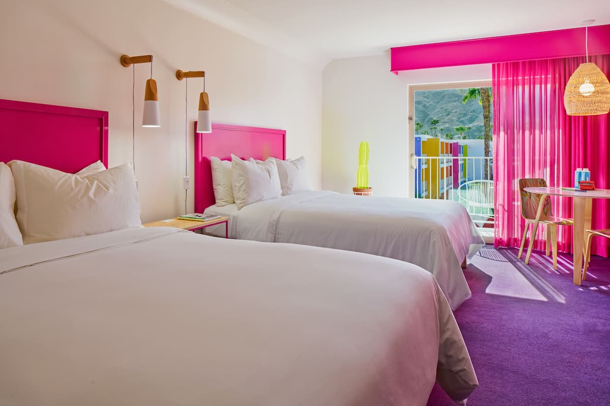 A hotel room with two beds, a desk with a chair, a window with pink curtains, and a view of a mountain.