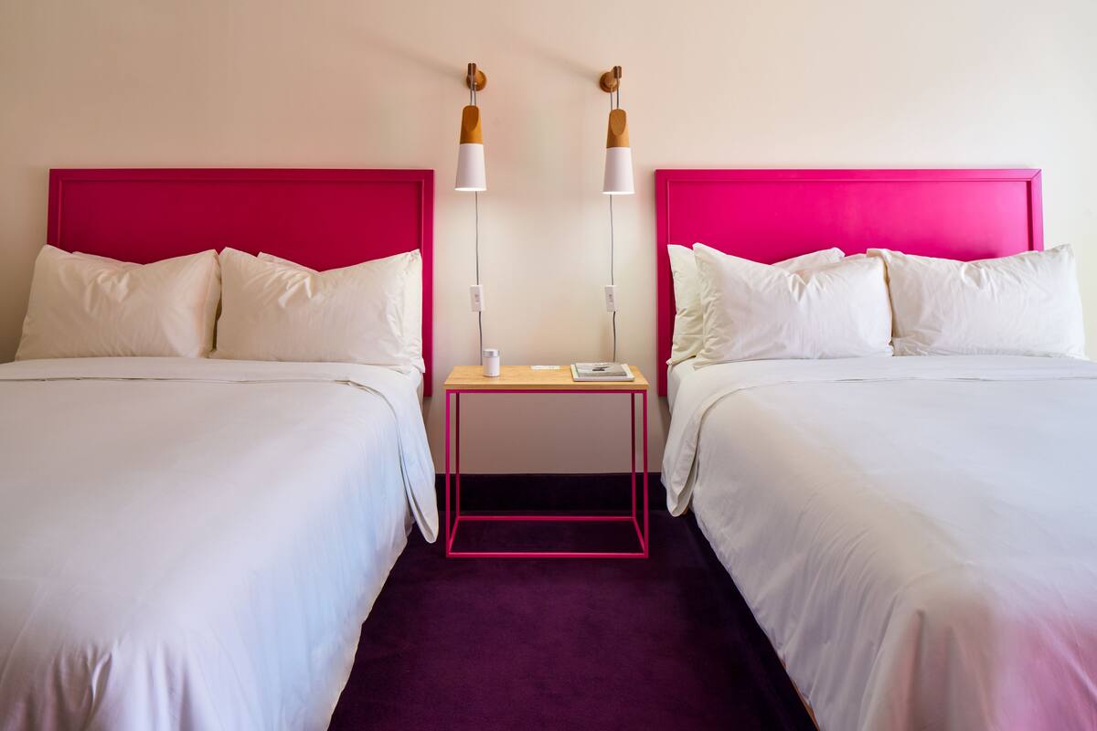 A hotel room with two beds, each with a pink headboard, white bedding, and a small wooden nightstand between them.