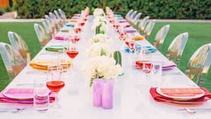 A long table set for a formal outdoor event with clear chairs, white tablecloth, and colorful table settings.