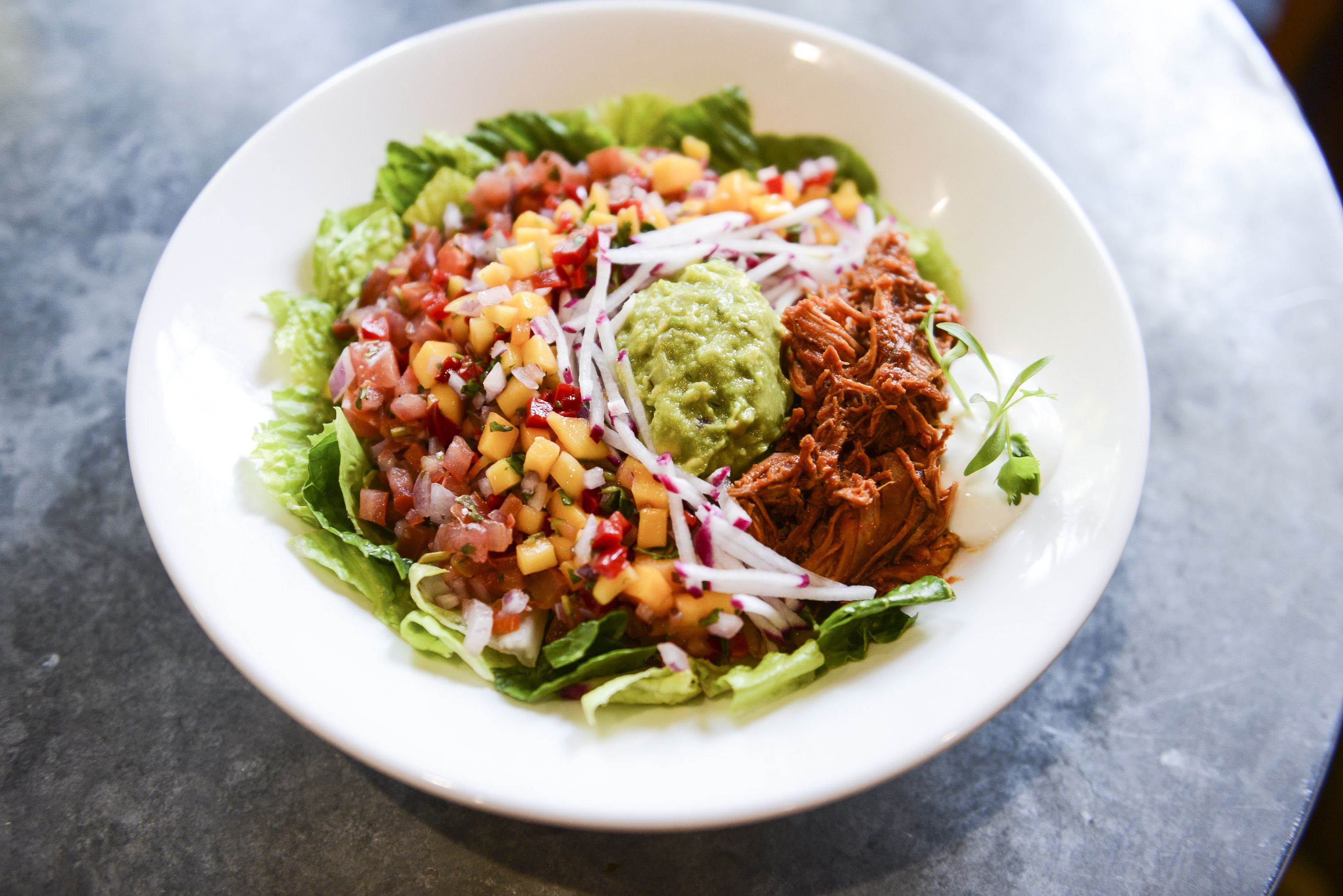 Eine Schüssel mit Essen, darunter zerrissenes Fleisch, gewürfelte Mango, Zwiebeln und Guacamole auf Salat.