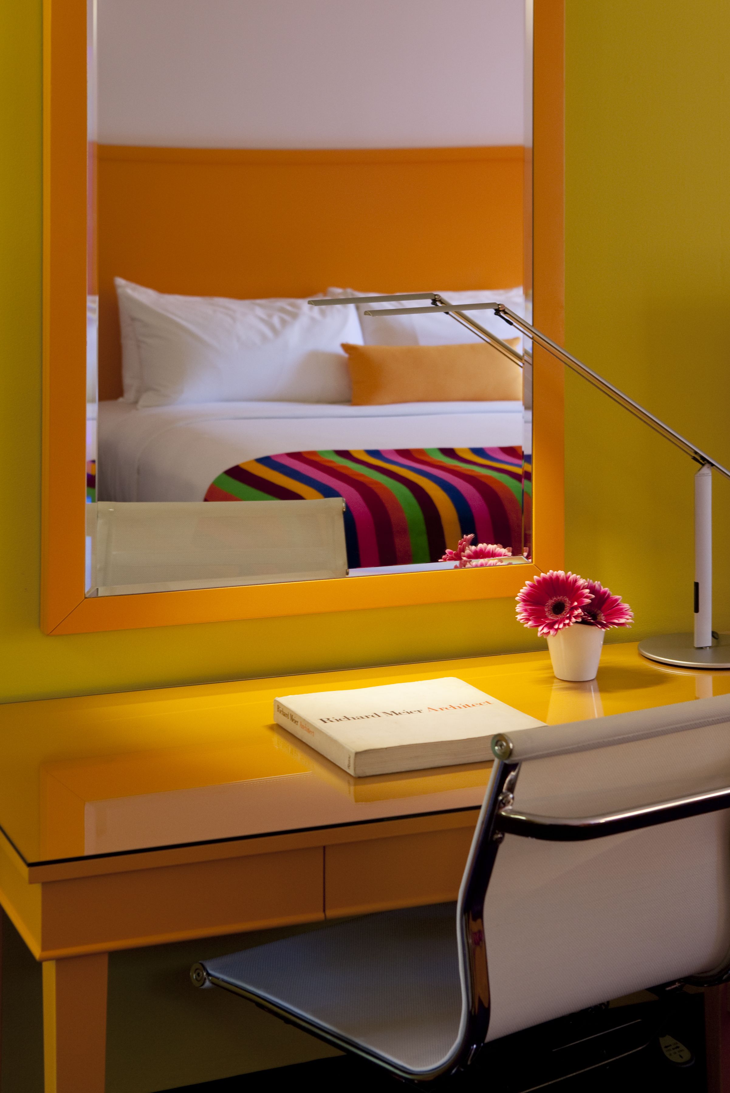 Una habitación de hotel con una cama, un escritorio con lámpara, una silla y un florero con flores.