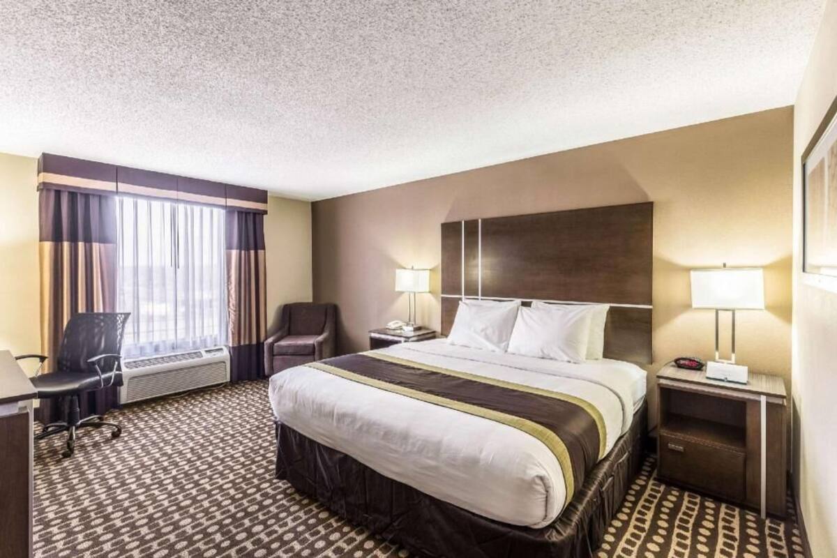A hotel room with a large bed, two bedside tables with lamps, a desk with a chair, a window with curtains, and a wall-mounted air conditioning unit.