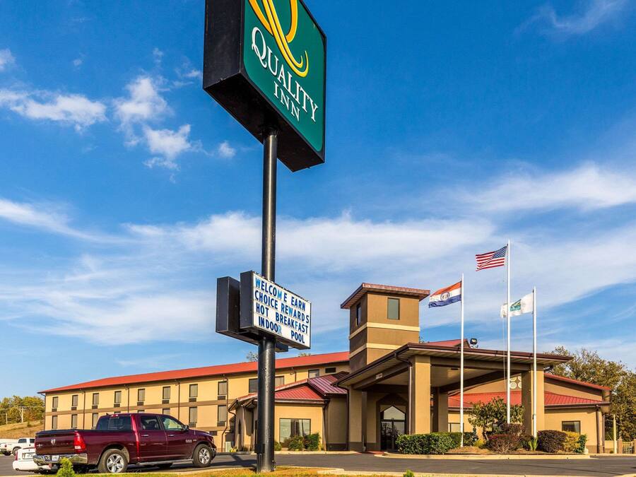 Een Quality Inn met een helderblauwe lucht, een donkerrode pickup geparkeerd ervoor, en twee vlaggen op het gebouw.
