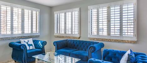 A living room with two blue sofas, a glass coffee table, and large windows with white shutters.
