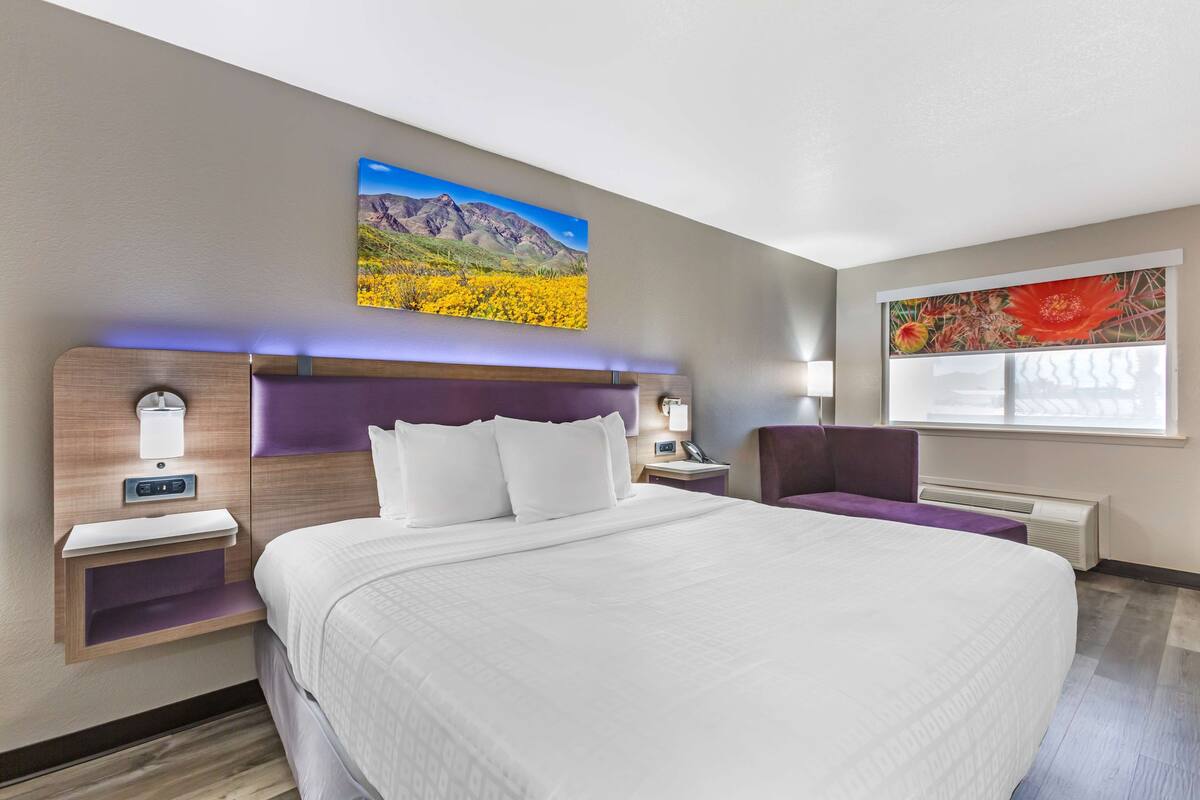 A modern hotel room with a large bed, a nightstand with a lamp, a chair, and two wall-mounted artworks.