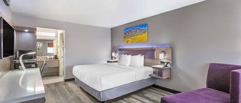 Una habitación de hotel moderna con una cama, un sillón morado, un escritorio con lavamanos y un cuadro en la pared.