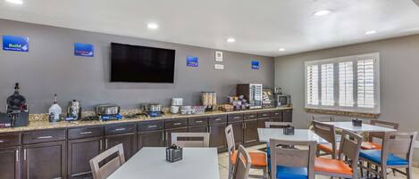 Een restaurantgedeelte in een hotel met meerdere tafels en stoelen, een keukenblok met diverse apparaten, en een grote flatscreen televisie aan de muur.