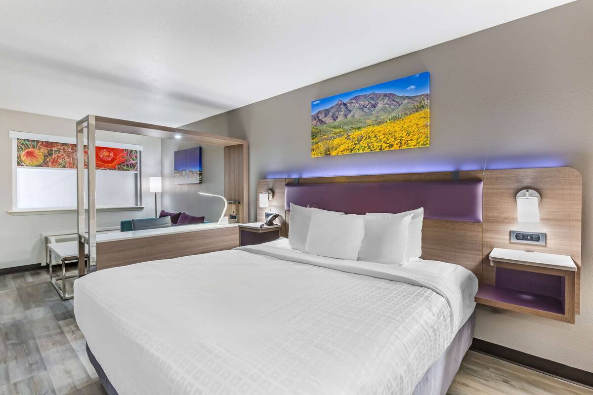 A modern hotel room with a large bed, a bedside table with a lamp, a nightstand, and a wall-mounted TV.