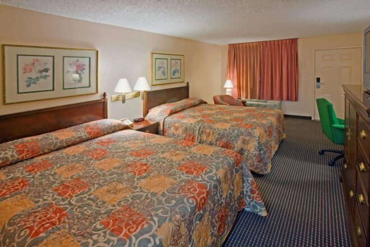 Ein Hotelzimmer mit zwei Betten, einem Schreibtisch, einem Stuhl, einer Kommode und gerahmten Bildern an den Wänden.