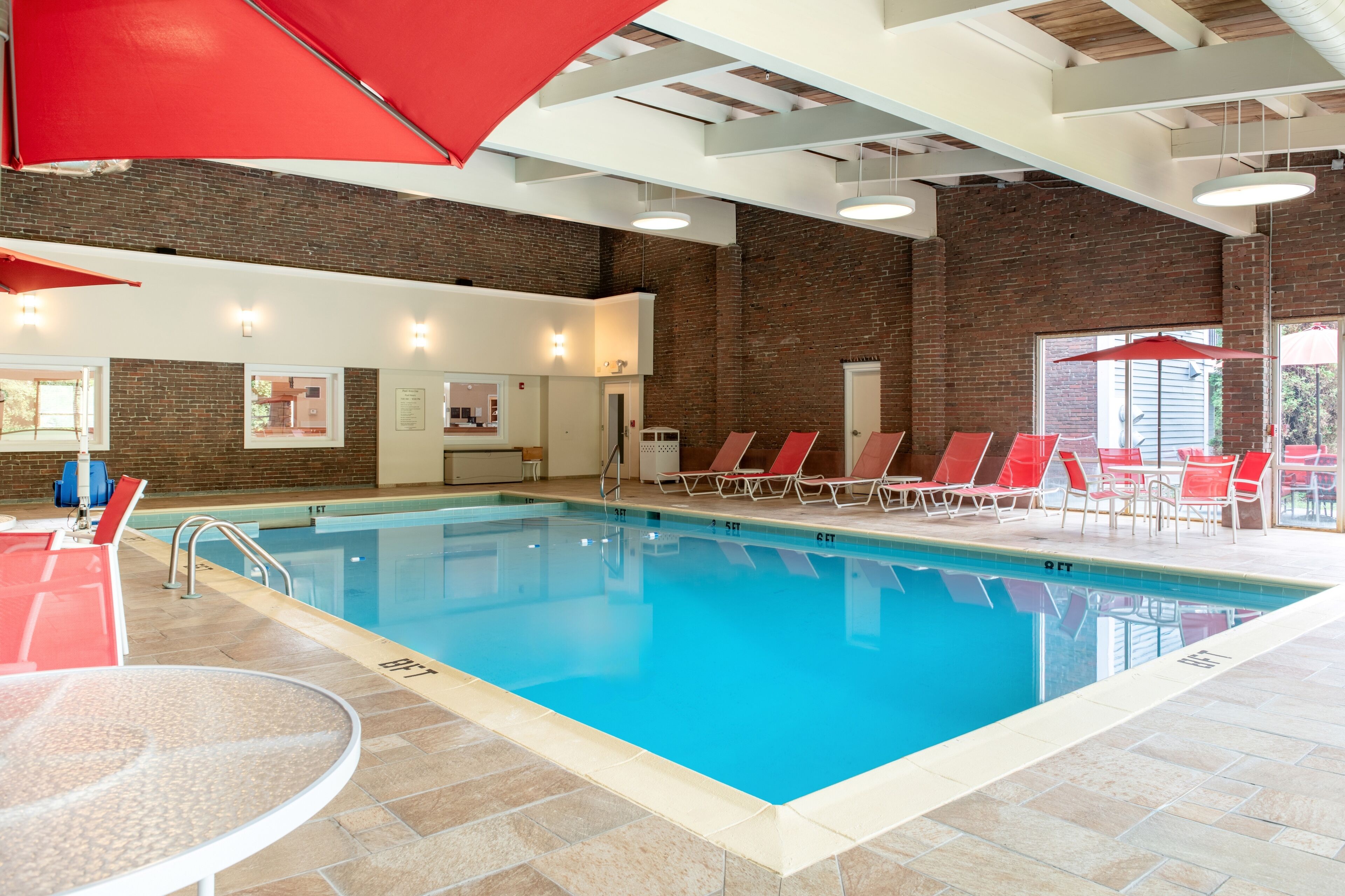 Une piscine intérieure à l’eau d’un bleu limpide, entourée de chaises et de parasols rouges, avec un mur de briques en arrière-plan.