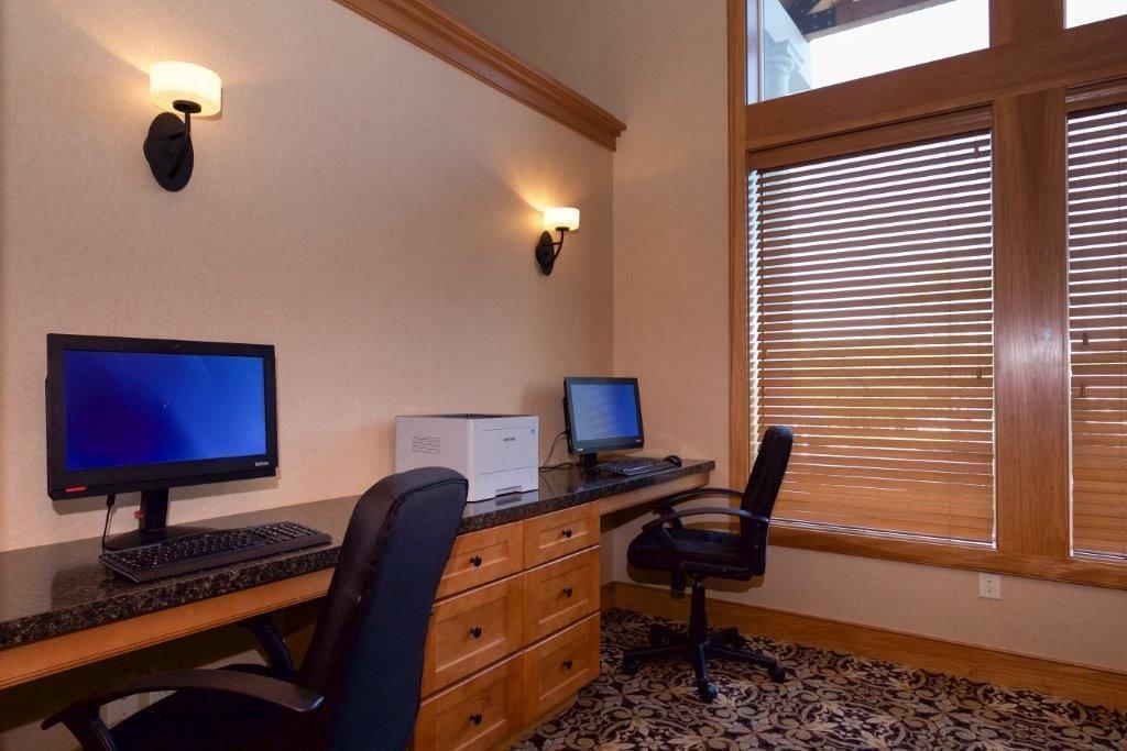 A room with two computer monitors, a printer, a desk, a chair, and a window with blinds.