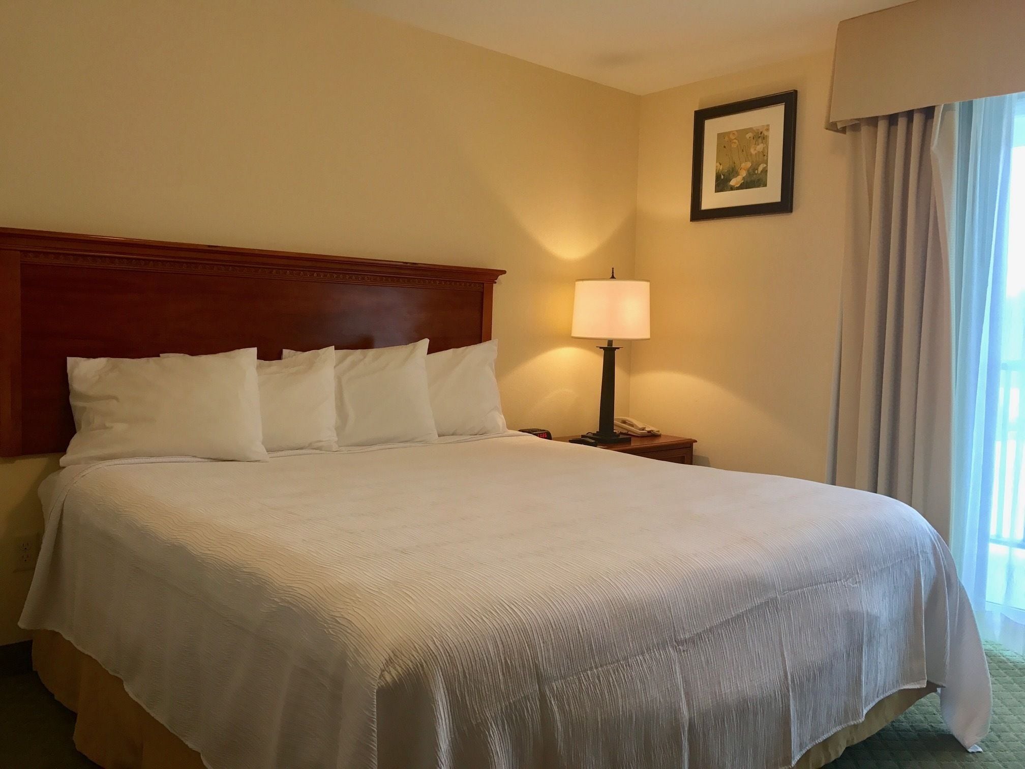 Una habitación de hotel con una cama grande, una mesita de noche con lámpara, un cuadro enmarcado en la pared y una ventana con cortinas.