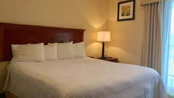 Una habitación de hotel con una cama grande, una mesita de noche con lámpara, un cuadro enmarcado en la pared y una ventana con cortinas.