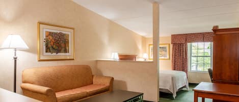 A hotel room with a sofa, a bed, a table, and a chair. There is a window with curtains, a lamp, and a framed picture on the wall.