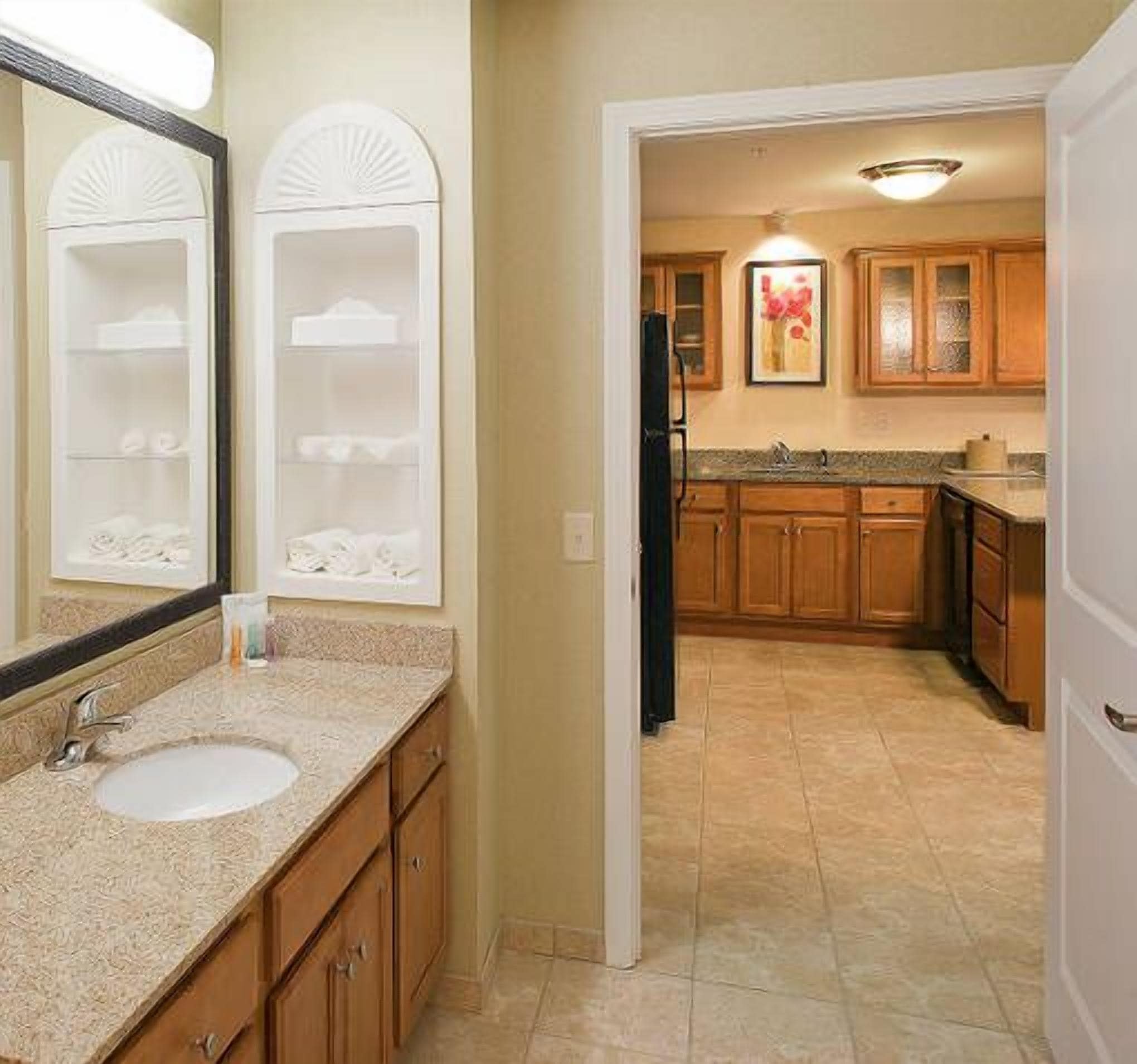 A bathroom with a sink, mirror, and two shelves. There is a doorway leading to a kitchen with wooden cabinets and a refrigerator.