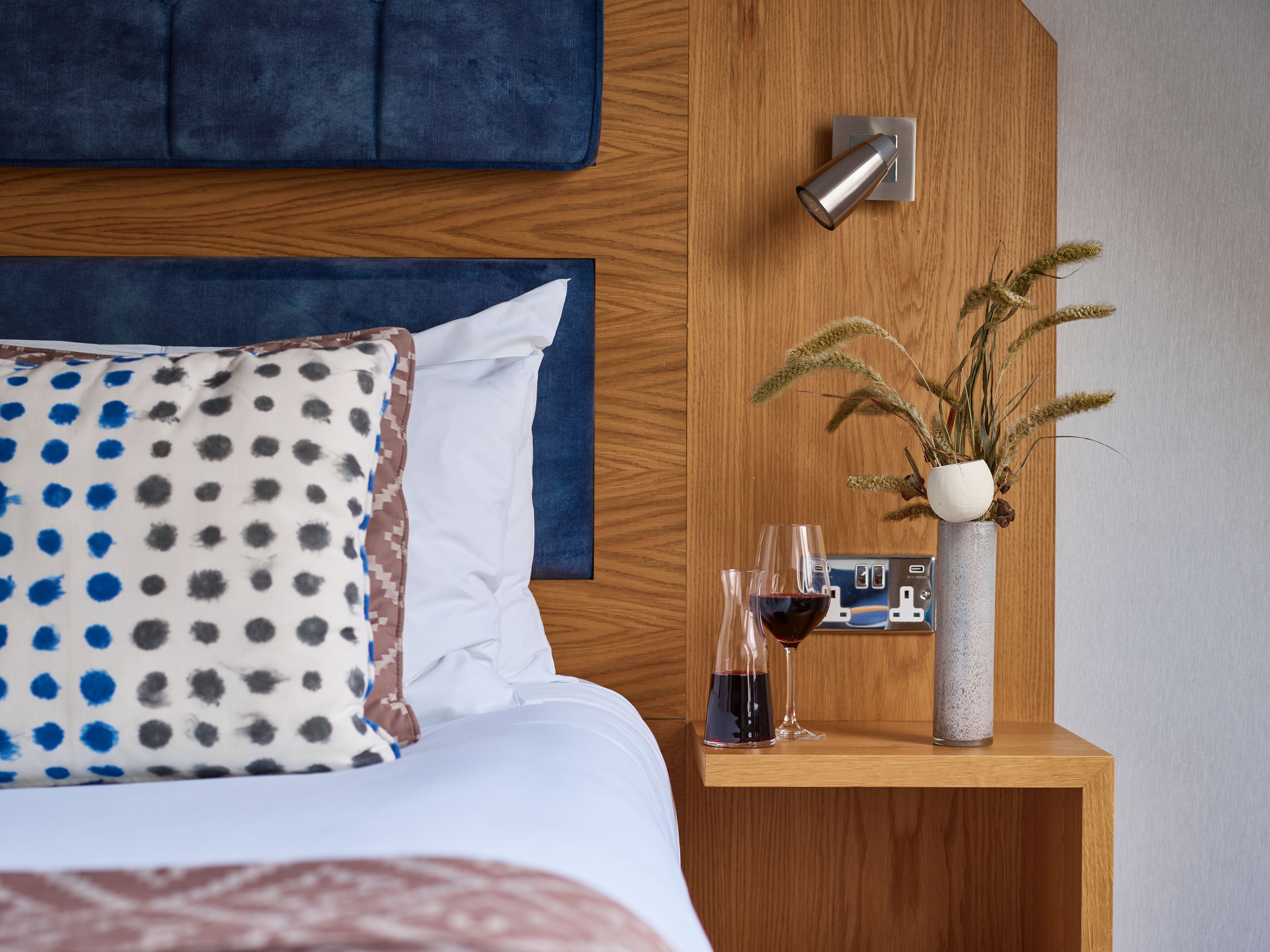 Ein ordentlich bezogenes Bett mit einem Kissen mit Punkten, ein Nachttisch aus Holz mit einer Vase mit Trockenpflanzen und zwei Gläsern Rotwein.