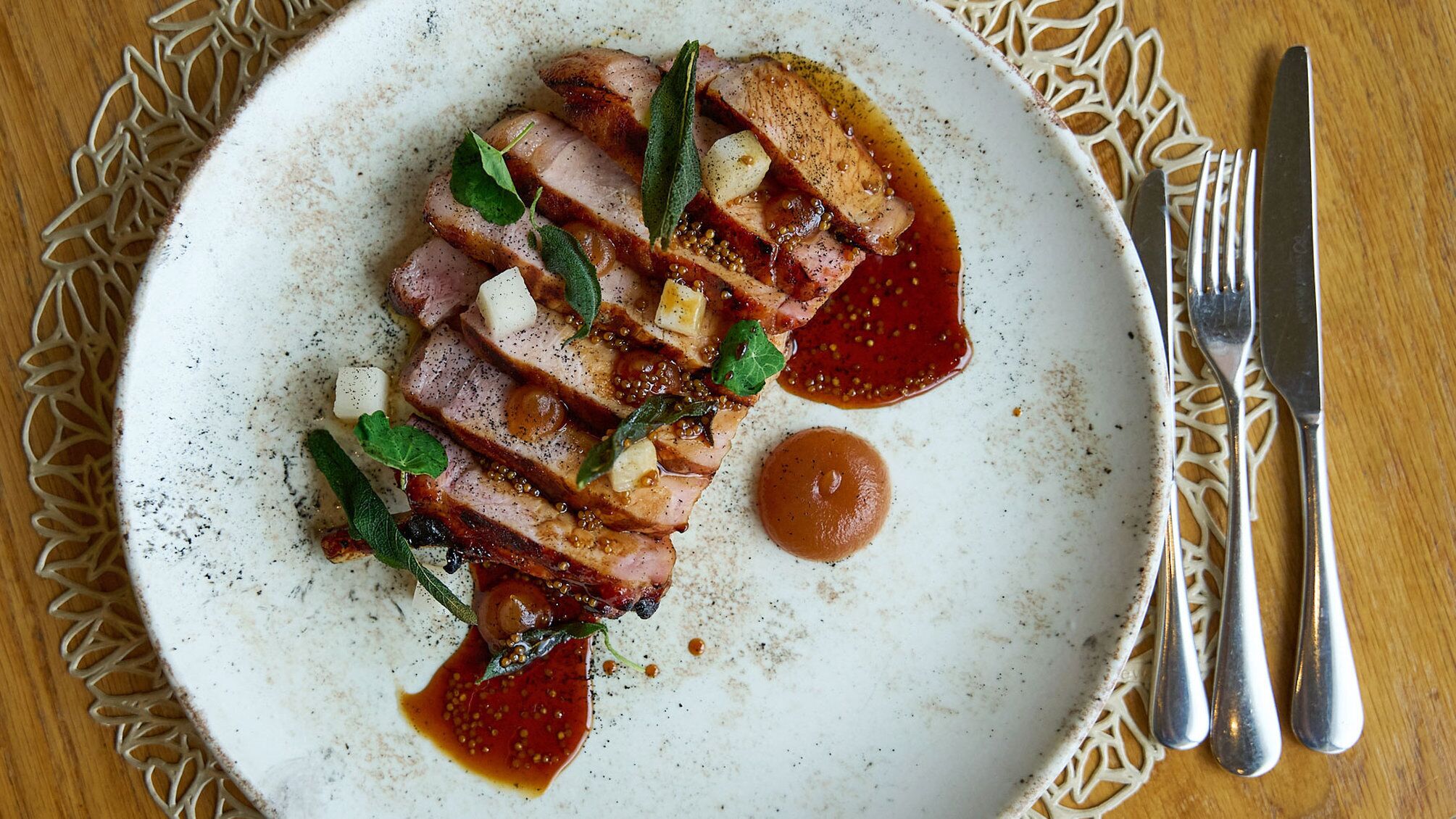 Un plato de carne en rodajas con salsa y decoración, acompañado de un tenedor y cuchillo sobre una mesa.