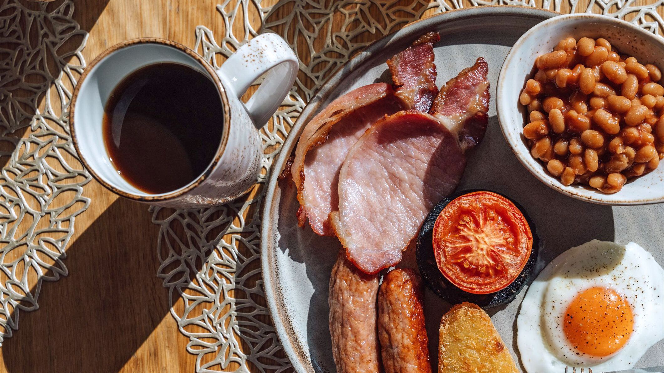 A breakfast tray with bacon, sausages, eggs, beans, and a tomato on a wooden table.